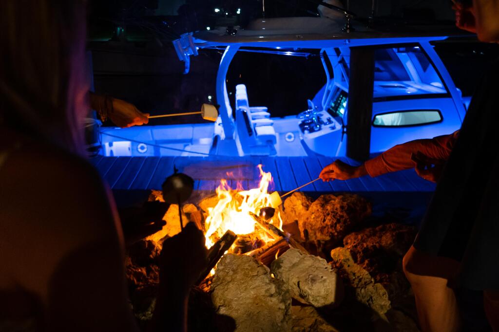 Nighttime beach gathering with illuminated boat and campfire, blue mood lighting highlighting the helm and cabin interior