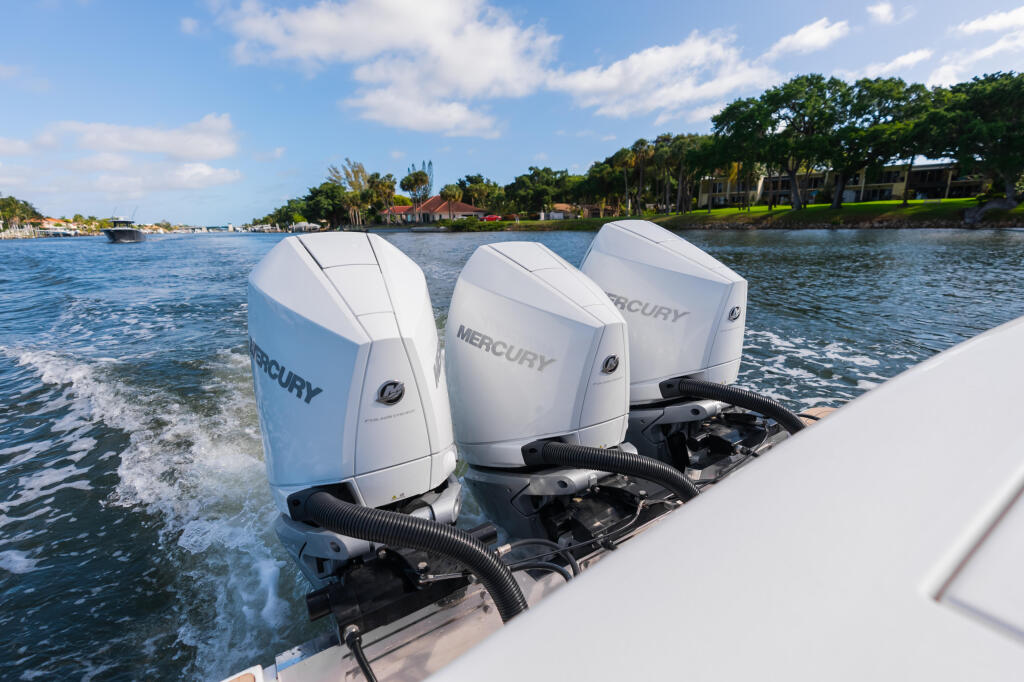 Three Mercury Verado outboard engines mounted on the transom of a boat at a riverside marina with waterfront homes in the background