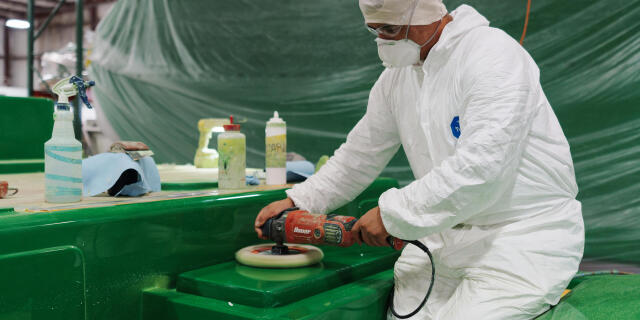 Worker in protective suit operating an orbital sander on a green fiberglass boat surface