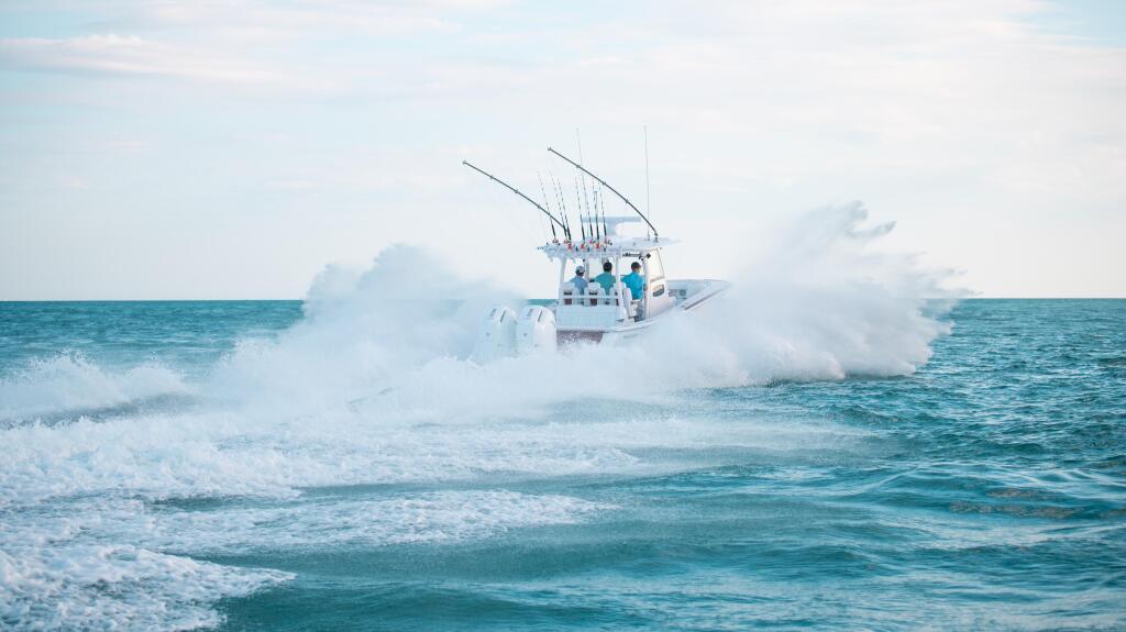 White sport fishing boat with multiple fishing rods riding through rough ocean waves near shore