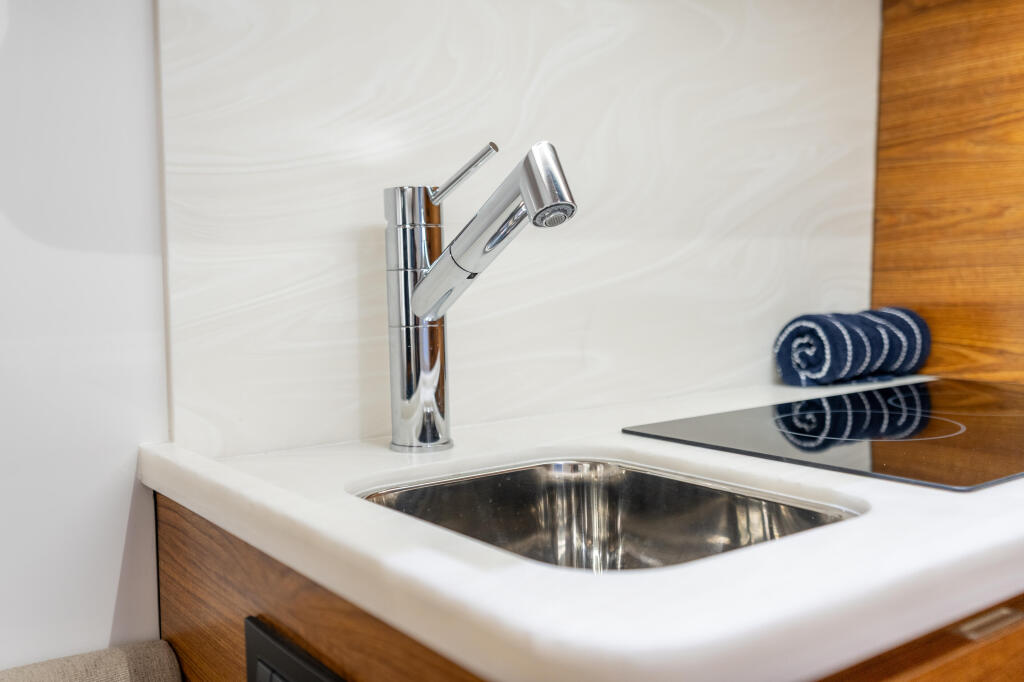 Modern galley sink with polished chrome faucet, stainless steel basin, and white Corian countertop with wood cabinetry