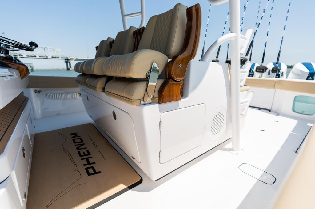 Leaning post with tan padded backrest and wood armrests in center of boat cockpit with fishing rods and helm visible