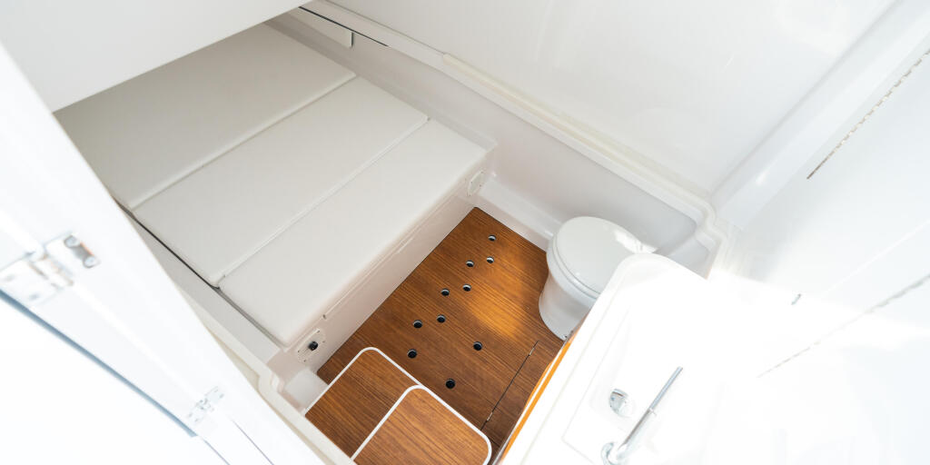Overhead view of a boat cabin with white cushioned seating, wooden flooring with drainage holes, and a white toilet fixture