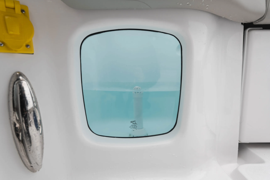 Cabin porthole window with tinted glass showing calm turquoise water and horizon, with steering wheel visible to the left