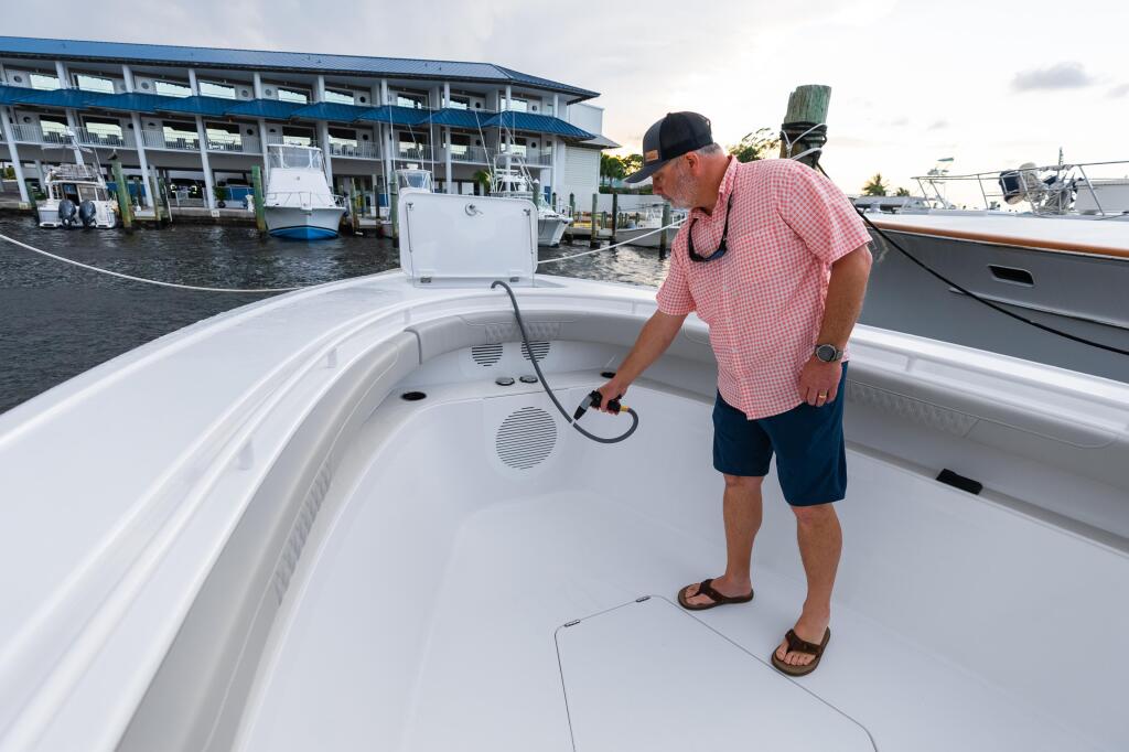 Man rinsing down a fishing boat's white console deck at a marina with multiple boat slips visible behind him