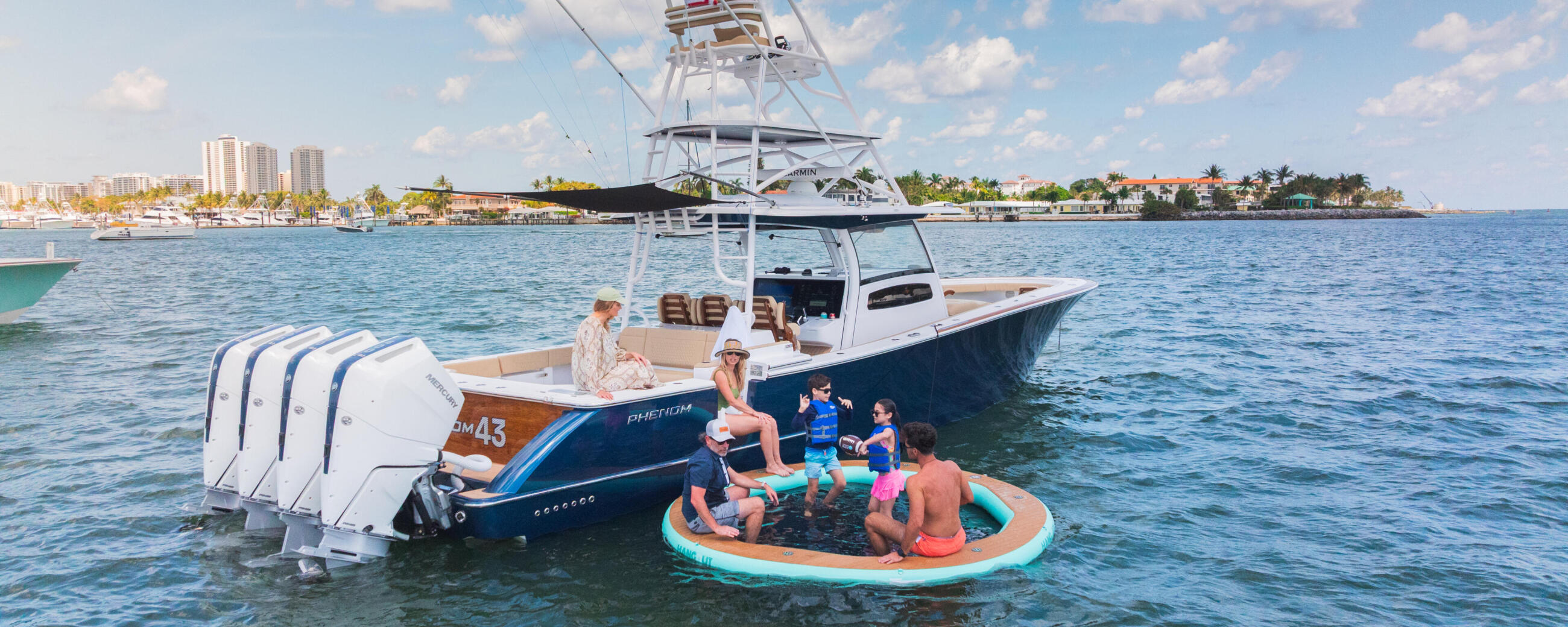 Family and children enjoying water activities on an inflatable raft beside a white and blue center console boat near a coastal shoreline