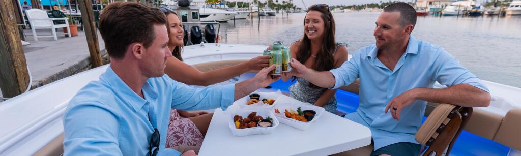 Four friends toast with drinks while dining at a table on a docked boat at a marina