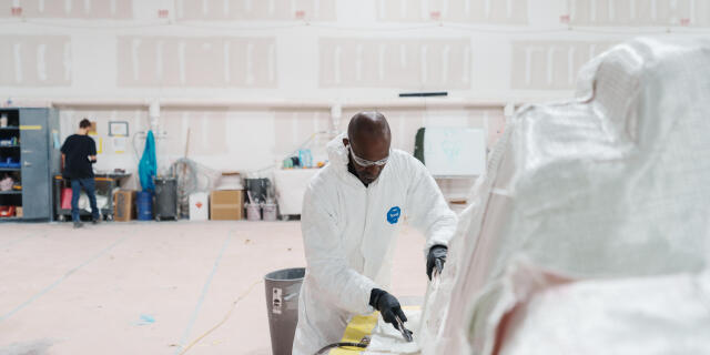 Worker in protective suit and gloves applying yellow tape to white fiberglass boat hull in industrial workshop