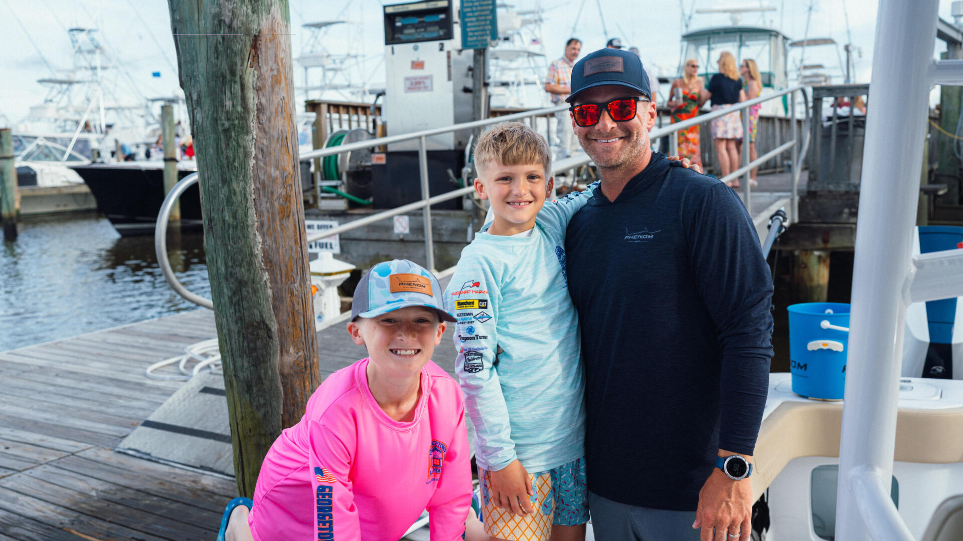 Father and two young children smile for the camera on a fishing boat dock surrounded by other vessels at a marina