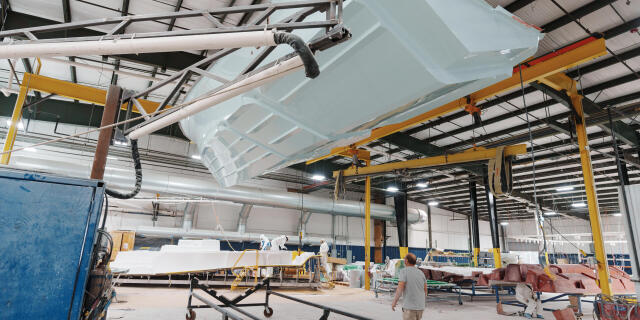Boat hull suspended from overhead crane in manufacturing facility with yellow support beams and work area below