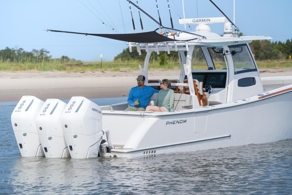Two people relax aboard a white center console fishing boat with triple outboard engines and extended hardtop canopy